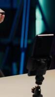 Vertical Gen Z girl in apartment studio using mobile phone to record, reviewing mini RGB lights. Young entertainer films electronics haul with cellphone on tripod, presenting LED lighting device, camera A video