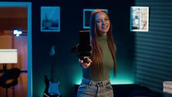 Young girl recording introductory with cellphone on selfie stick, inviting viewers to subscribe to channel. Media star in home studio uses mobile phone to film herself, camera A video