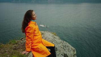 Happy Brunette Woman In Orange Raincoat Sitting At Top Of Rock On Ocean Coastline, Enjoying Solitude video