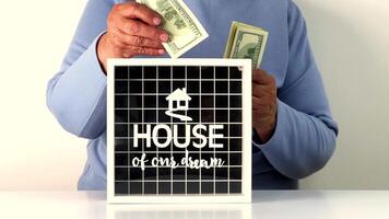 A person is actively saving money by inserting cash into a savings box labeled House of Our Dream. This symbolizes dedication towards homeownership aspirations video