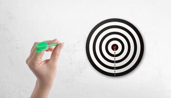 Hand holding a dart aiming at a black-and-white dartboard on a wall, representing focus, precision, and achieving goals in a dynamic setup photo