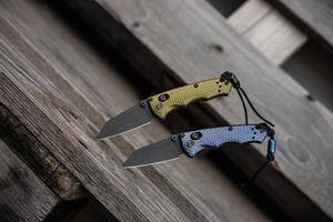 Two folding knives with textured handles, one green and one blue, are displayed on a rustic wooden surface photo