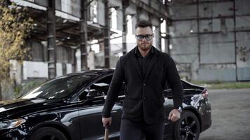 Strong man with a bat in abandoned place. Stylish man in glasses and black wearing moving forward on expensive car background inside the old empty plant. video