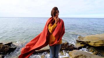 Hiker woman wearing yellow sweater trekking in cold sunny weather, hair blowing in wind. Portrait of a lady wrapped in a blanket on beach in autumn video