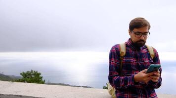 man is sitting on the road of a mountain and texting a message on his smartphone. He has a large backpack on his shoulders. Hiking in the mountains. video