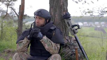 Man in a military uniform and a bulletproof vest sits in the forest near a metal detector and a military backpack. A man pauses in the work of demining the territory video