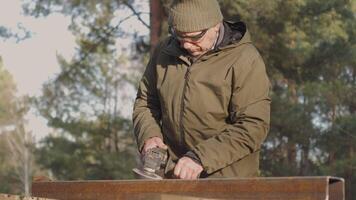 A man in a warm hat and jacket is using a power tool to shape metal in a forested area, showcasing craftsmanship amidst nature during daytime. video