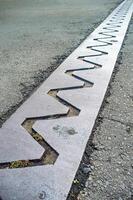 A close up view of a bridge expansion joint with a zigzag pattern, designed to accommodate structural movements and prevent damage. The metal plate is embedded in the asphalt road surface photo