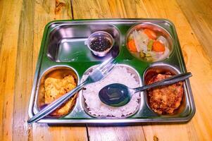 A nutritious school lunch featuring rice, fried egg, vegetables, and a dipping sauce, served in a stainless steel tray with a fork and spoon on a wooden table photo