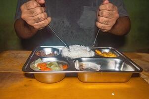 A person is holding a fork and spoon, ready to eat a nutritious meal consisting of rice, vegetables, and protein, served in a stainless steel tray, symbolizing a healthy and balanced diet photo