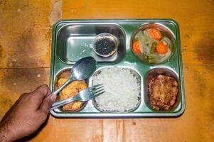 A stainless steel lunch tray with a nutritious meal consisting of rice, fried fish, an omelette, and a small container of sauce. A hand is holding a spoon and fork, ready to eat photo