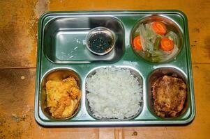 A nutritious and balanced free lunch featuring rice, fried chicken, vegetable soup, and a dipping sauce, served in a stainless steel tray on a rustic wooden table photo