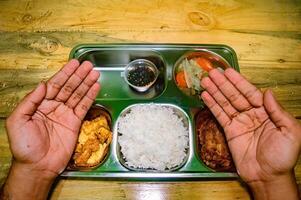 A pair of hands shown in a prayer-like gesture over a metal tray containing a balanced meal with rice, vegetables, fried food, and a small cup of sauce, symbolizing gratitude for a nutritious lunch photo