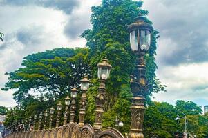 A row of ornate street lamps with intricate designs lines a pathway, set against a backdrop of lush green trees and a cloudy sky, creating a picturesque and historical ambiance photo