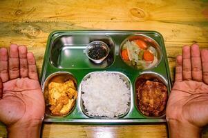 A stainless steel tray with a balanced meal including rice, fried food, vegetables, and sauce, with hands on either side, symbolizing a healthy and nutritious free lunch concept photo
