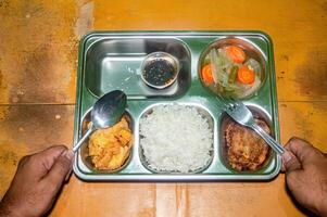 A nutritious and balanced free lunch served on a metal tray, featuring rice, fried chicken, vegetables, and a dipping sauce. Hands holding a spoon and fork are ready to enjoy the meal photo