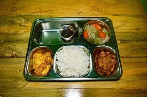 A nutritious and healthy free lunch concept featuring a stainless steel tray with rice, fried chicken, vegetables, and a dipping sauce, placed on a rustic wooden table photo