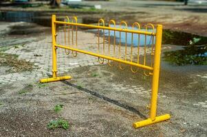 A yellow metal barricade stands on a cracked, weathered pavement with puddles and sparse grass, indicating an outdoor area possibly under maintenance or restricted access photo