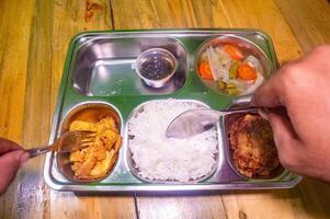 A stainless steel lunch tray with rice, fried tofu, vegetables, and a fried patty. Hands are holding a fork and spoon, ready to eat. Ideal for concepts of free or nutritious meals photo