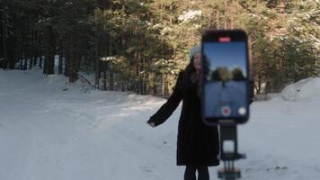 Young woman in winter attire dancing playfully among snow covered trees, recording herself with smartphone mounted on tripod in scenic forest landscape video
