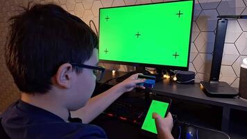 A dark- haired boy scroll a smartphone and holds a bank card, sitting in front of a monitor video