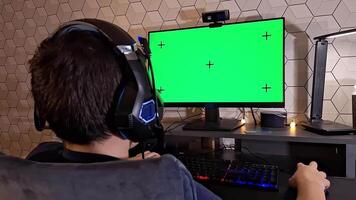 Rear view of boy uses a computer, sits with headphones in front of a green monitor screen video