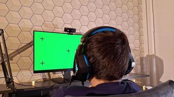 Rear view of boy uses a computer, sits with headphones in front of a green monitor screen video