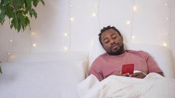 Sleepy african american guy browsing internet on smartphone before sleeping. video