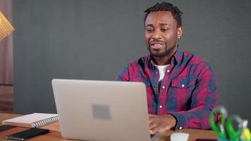joyful black man chatting by call in laptop from his home office, smiling to web camera video