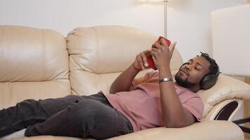 Resting african american guy in headphones wathcing browsing on phone. video