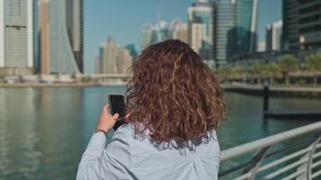 Rizado mujer tomando foto por teléfono inteligente de increíble edificio en dubai en ciudad centro, espalda ver video