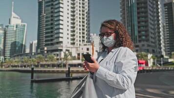 Tourist woman in medical face mask browsing phone walking on waterfront in city. video
