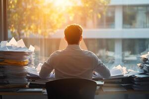 A man in a white shirt works works at an office desk filled with stacks of business papers and documents, in front of a large panoramic window. Business and office life. photo