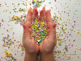 Child playing with colored sugar candies on white background. Confectionery sprinkles in child's palms. Children's game, fine motor skills, mess. photo