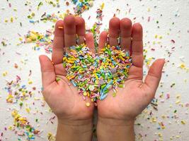 Child's hands are covered with multi-colored sugar sprinkles and form a heart shape. Child plays with small sprinkles topping. Mother's Day. photo