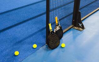 A padel or paddle tennis racket and a bright yellow tennis ball lie on a blue court. The net and a blurred figure of a player are visible in the background. photo