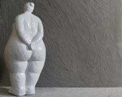 A white plaster figurine of a plump woman turned away from us, set against a gray background with copy space in a minimalist style photo