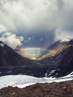 scenery with bright rainbow above glacier in mountain valley. Top view to colorful rainbow and low clouds in mountains in rainy and sunny weather. photo