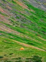 Tourist tent far away on a green mountain slope. Minimalistic landscape of a green mountain slope with a small tent. Scenic alpine landscape with tent on green hill among rocks. photo