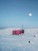 Lonely red house on a snowy frosty hill under evening sky with a full moon. Vertical minimalistic background with of lonely red cabin in winter. photo