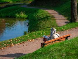A woman in a headscarf is sitting on a bench in an autumn park and leafing through a magazine. Outdoor recreation photo