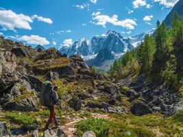 Solo hiking with a large backpack in the mountains. A man climbs a rocky mountain slope. In the background is a dome of snow-capped mountains under a blue cloudy sky. Lonely man walking in a mountain. photo