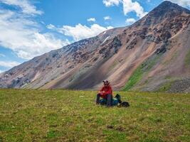 The dreaming hiker sits on a large backpack and happily looks into the distance against the background of blue dramatic clouds against the background of tundra mountains. Rest time on a hike photo