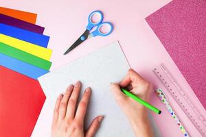 Woman's hand holds a pencil and draws a hearts on paper on pink background. DIY concept for Valentine's day card crafts. Top view. Flat lay. Step by step photo instruction. Step 2.