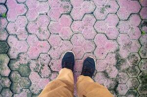A person wearing black shoes and brown pants standing on a pink hexagonal tiled pavement with some moss and dirt. This image highlights casual urban fashion and unique pavement design photo