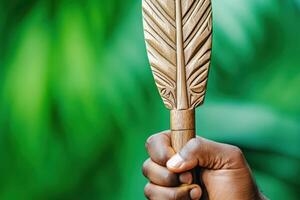 Close-up of a hand gripping a traditional wooden spear adorned with intricate carvings and vibrant feathers against a blurred green background photo