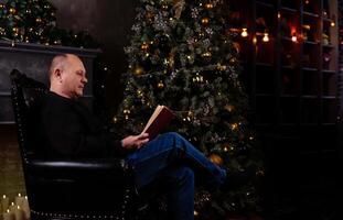 man sits comfortably in a chair in the living room, engrossed in a book while the festive Christmas tree glows nearby, creating a cozy atmosphere photo