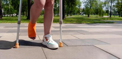 leg encased in an orange cast, with crutches visible in the lush, green background of a summer park setting. Concept of injury recovery photo