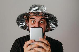 Man with shocked expression wearing tin foil hat holding smartphone photo