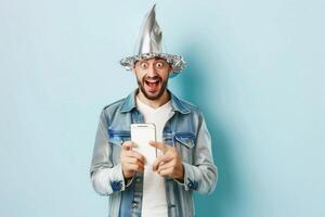 Excited man in tin foil hat using smartphone against blue background photo